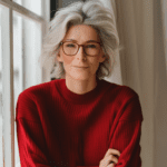 
A woman in her 40s with stylish silver hair and red glasses, smiling in a red knit sweater, by a window with soft light.